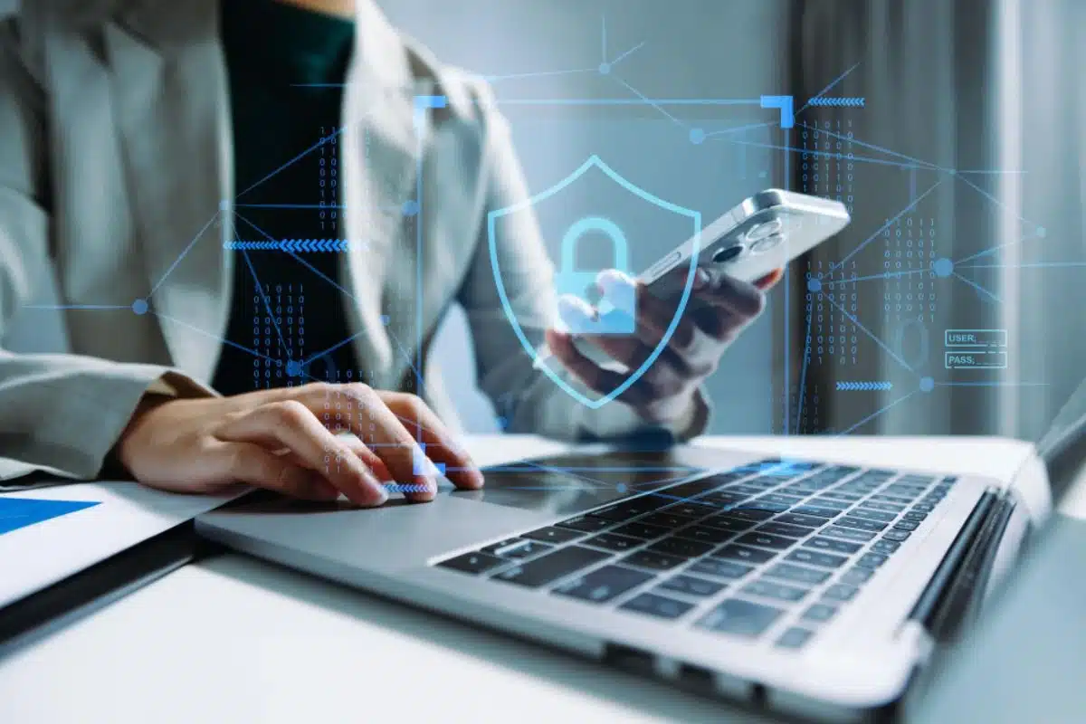 Executivo mexendo no celular com o notebook aberto, representando a segurança digital.